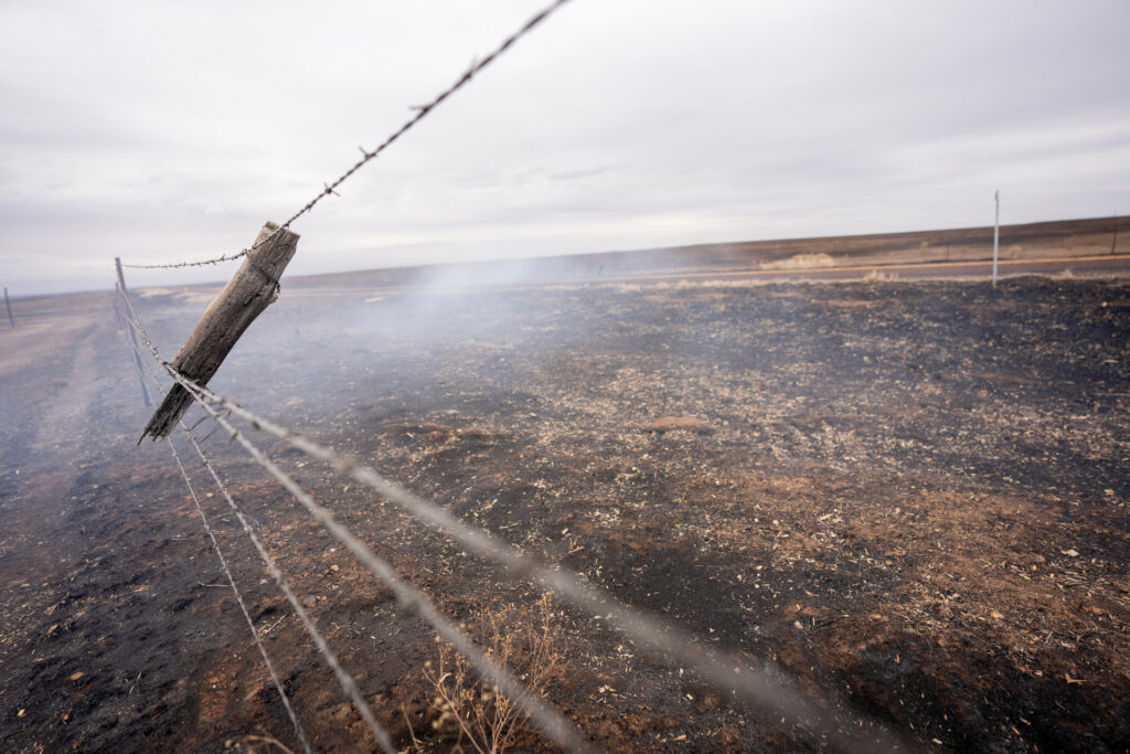 The top portion of a wooden fence post swings from two strands of barbed wire and still smolders