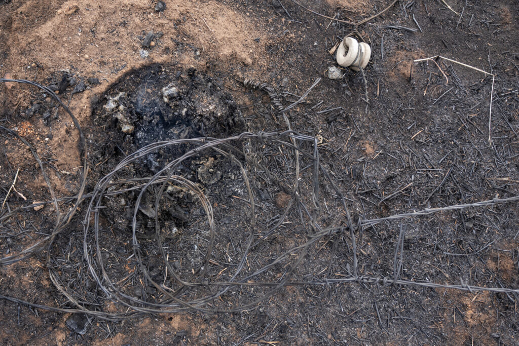 barbed wire lies coiled up around the remnants of a burned out fence post on charred ground.