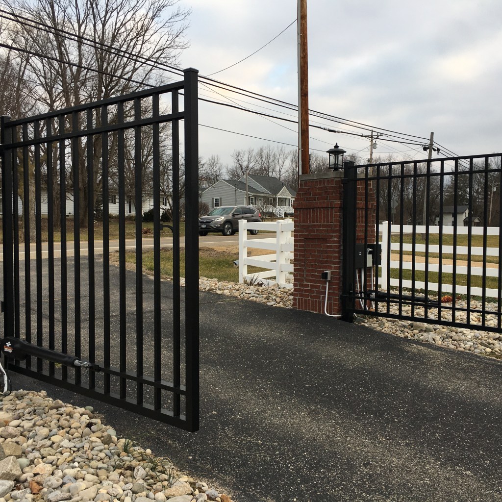 A black metal driveway gate with brick pillars, opening onto a paved road. A black metal driveway gate with brick pillars, opening onto a paved road.