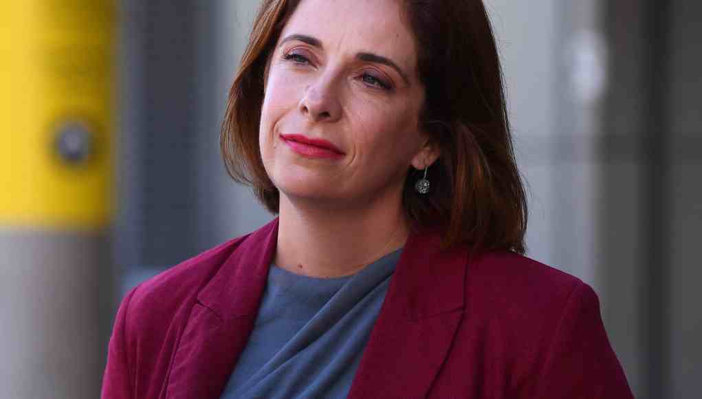 Mujer de cabello castaño medio, ligeramente ondulado, vestida con una chaqueta burdeos y una blusa gris azulada. Está de pie en un entorno urbano, con expresión serena o reflexiva. Al fondo se ven estructuras modernas desenfocadas, incluyendo una columna amarilla. Mujer de cabello castaño medio, ligeramente ondulado, vestida con una chaqueta burdeos y una blusa gris azulada. Está de pie en un entorno urbano, con expresión serena o reflexiva. Al fondo se ven estructuras modernas desenfocadas, incluyendo una columna amarilla.