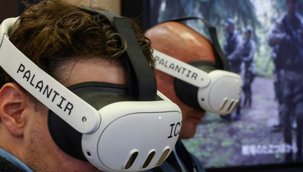 Dos personas con cascos de realidad virtual personalizados con la marca "Palantir", utilizados en una demostración tecnológica. Al fondo, se muestra una pantalla con una imagen de soldados en un entorno forestal, lo que sugiere un contexto de simulación militar o entrenamiento táctico en entornos virtuales. Dos personas con cascos de realidad virtual personalizados con la marca "Palantir", utilizados en una demostración tecnológica. Al fondo, se muestra una pantalla con una imagen de soldados en un entorno forestal, lo que sugiere un contexto de simulación militar o entrenamiento táctico en entornos virtuales.