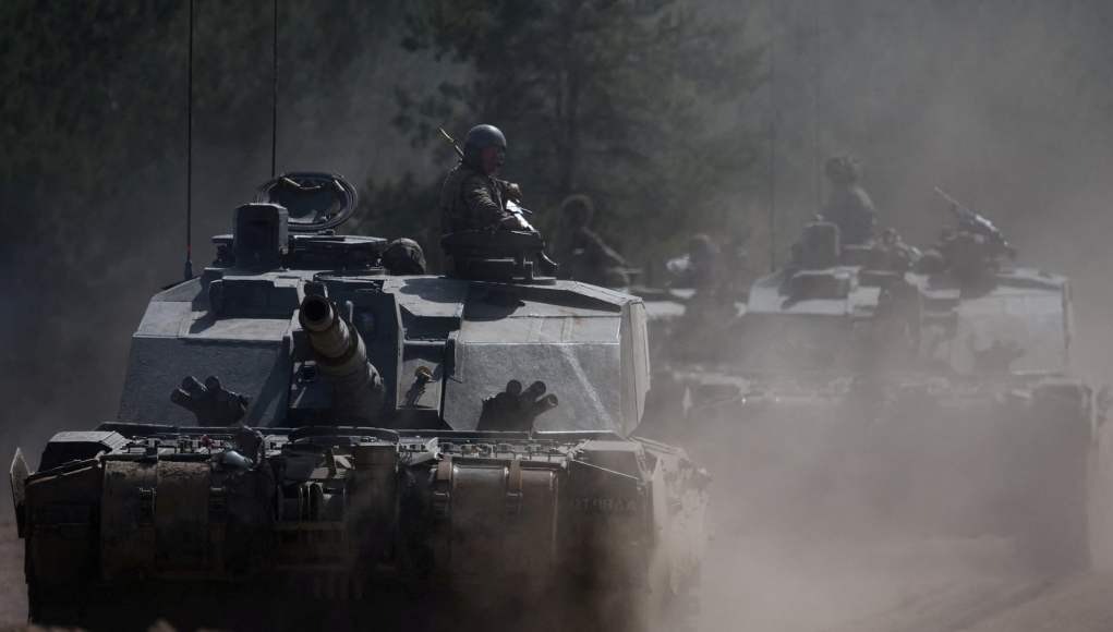 Columna de tanques militares avanzando por un terreno polvoriento rodeado de árboles, con soldados visibles sobre los vehículos mientras levantan nubes de polvo a su paso. Columna de tanques militares avanzando por un terreno polvoriento rodeado de árboles, con soldados visibles sobre los vehículos mientras levantan nubes de polvo a su paso.