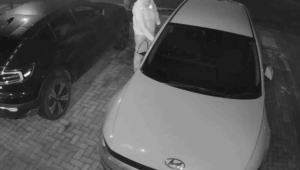Captura nocturna de una cámara de seguridad que muestra a una persona encapuchada y con el rostro cubierto intentando abrir la puerta de un coche Hyundai blanco estacionado en una entrada residencial, junto a otro coche negro. Captura nocturna de una cámara de seguridad que muestra a una persona encapuchada y con el rostro cubierto intentando abrir la puerta de un coche Hyundai blanco estacionado en una entrada residencial, junto a otro coche negro.