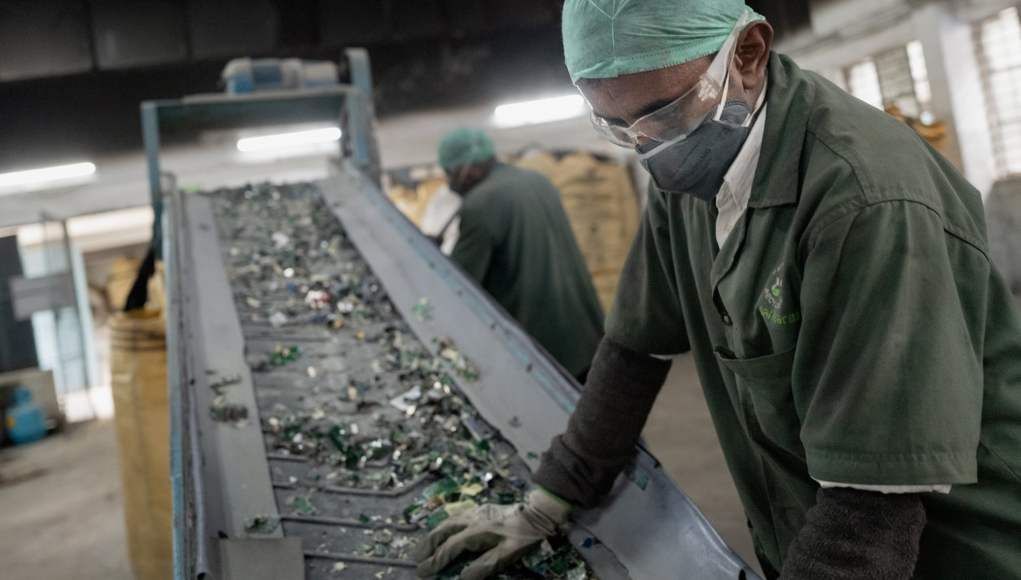 Trabajador con uniforme verde, guantes, mascarilla y gorro de protección, opera una cinta transportadora que lleva desechos electrónicos en una planta de reciclaje. En el fondo, otro trabajador realiza labores similares en un ambiente industrial iluminado. Trabajador con uniforme verde, guantes, mascarilla y gorro de protección, opera una cinta transportadora que lleva desechos electrónicos en una planta de reciclaje. En el fondo, otro trabajador realiza labores similares en un ambiente industrial iluminado.