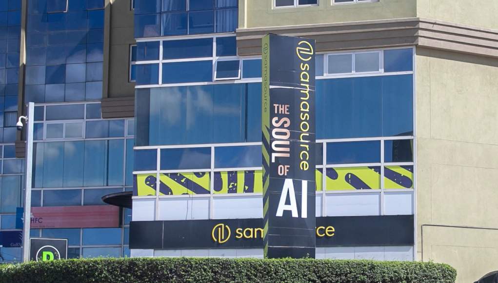 Edificio con fachada de vidrio azul y paredes verdes, con un letrero vertical que dice "Samasource The Soul of AI" en letras grandes y destacadas. Abajo, se ve el logo y el nombre "Samasource" en amarillo sobre fondo negro. El área exterior tiene arbustos verdes y una cámara de seguridad montada en un poste. Edificio con fachada de vidrio azul y paredes verdes, con un letrero vertical que dice "Samasource The Soul of AI" en letras grandes y destacadas. Abajo, se ve el logo y el nombre "Samasource" en amarillo sobre fondo negro. El área exterior tiene arbustos verdes y una cámara de seguridad montada en un poste.