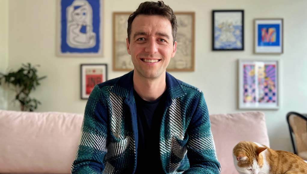 Imagen de un hombre joven sonriendo, sentado en un sofá de color claro en un ambiente hogareño. Lleva una chaqueta de cuadros en tonos verdes y azules sobre una camiseta negra. Detrás de él, se pueden ver varias obras de arte colgadas en la pared, con marcos de diferentes tamaños y estilos. A la derecha, un gato de color naranja y blanco está sentado en el sofá, mirando hacia abajo. En la esquina izquierda, hay una planta en el fondo, añadiendo un toque de verde al entorno. Imagen de un hombre joven sonriendo, sentado en un sofá de color claro en un ambiente hogareño. Lleva una chaqueta de cuadros en tonos verdes y azules sobre una camiseta negra. Detrás de él, se pueden ver varias obras de arte colgadas en la pared, con marcos de diferentes tamaños y estilos. A la derecha, un gato de color naranja y blanco está sentado en el sofá, mirando hacia abajo. En la esquina izquierda, hay una planta en el fondo, añadiendo un toque de verde al entorno.