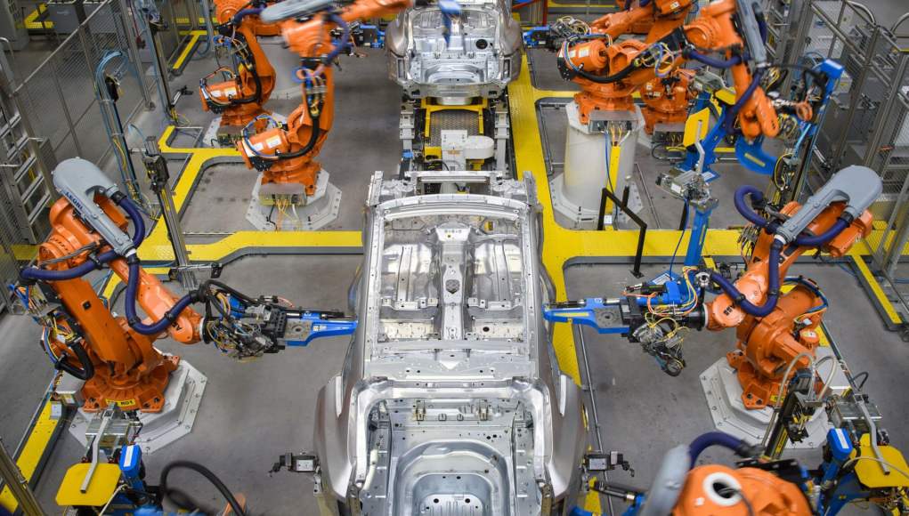 Línea de producción automatizada en una fábrica de automóviles, con múltiples brazos robóticos naranjas ensamblando carrocerías metálicas de coches. Línea de producción automatizada en una fábrica de automóviles, con múltiples brazos robóticos naranjas ensamblando carrocerías metálicas de coches.