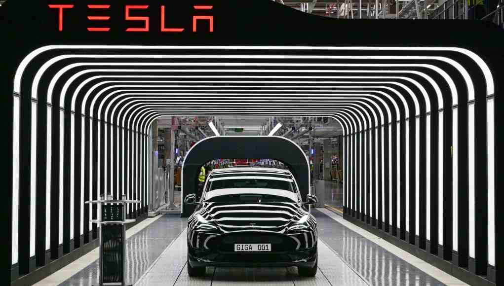 Automóvil Tesla negro saliendo de una línea de producción en una fábrica. El vehículo atraviesa un túnel iluminado con franjas LED blancas que crean un efecto visual futurista. Sobre la estructura del túnel, se ve el logotipo de Tesla en letras rojas. La matrícula del coche dice "GIGA 001", indicando su fabricación en la Gigafactory. Automóvil Tesla negro saliendo de una línea de producción en una fábrica. El vehículo atraviesa un túnel iluminado con franjas LED blancas que crean un efecto visual futurista. Sobre la estructura del túnel, se ve el logotipo de Tesla en letras rojas. La matrícula del coche dice "GIGA 001", indicando su fabricación en la Gigafactory.