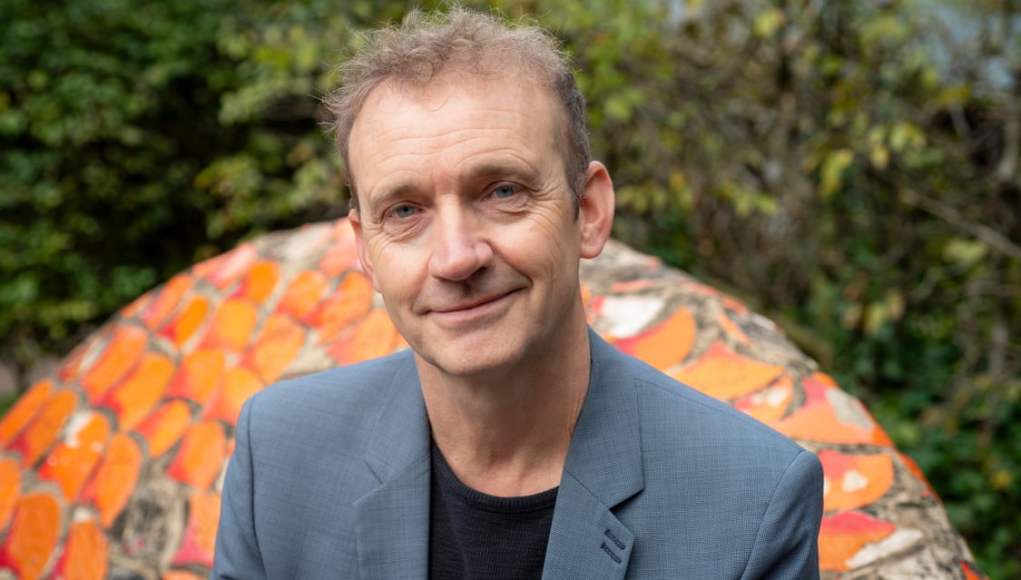 Un hombre de mediana edad con cabello corto y rizado, vistiendo una chaqueta gris y una camiseta negra, sonríe levemente mientras posa al aire libre. De fondo se puede apreciar un mosaico colorido con tonos naranjas y un entorno natural con vegetación difusa. Un hombre de mediana edad con cabello corto y rizado, vistiendo una chaqueta gris y una camiseta negra, sonríe levemente mientras posa al aire libre. De fondo se puede apreciar un mosaico colorido con tonos naranjas y un entorno natural con vegetación difusa.