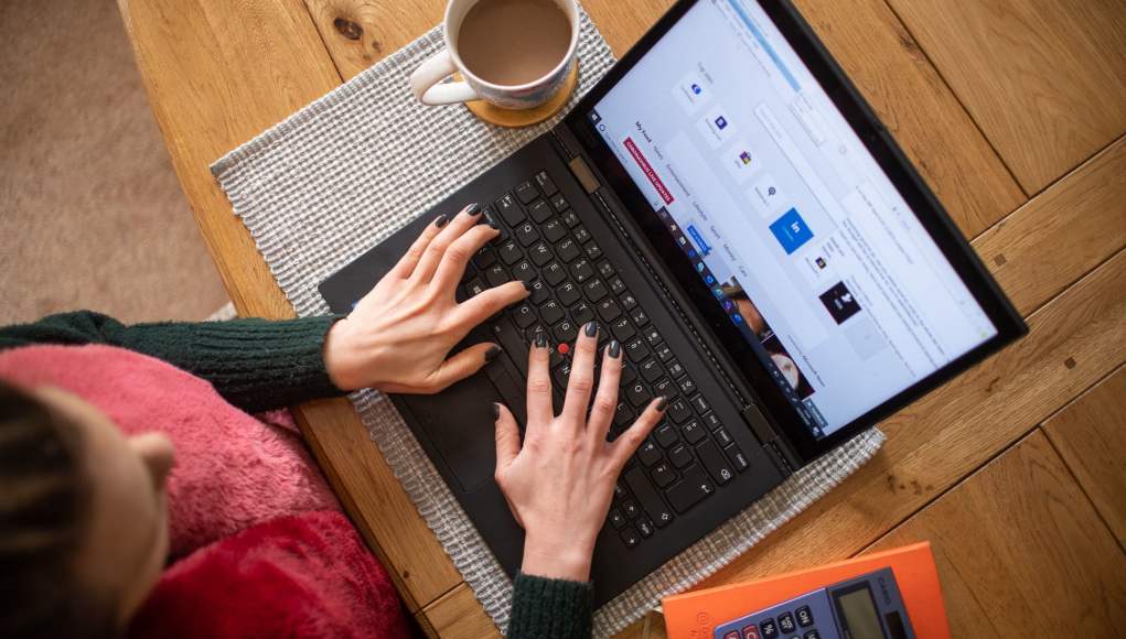 Una vista desde arriba de una persona trabajando en una computadora portátil. Se pueden ver las manos de la persona tecleando en el teclado del portátil. La computadora está colocada sobre una tela de cuadros en una superficie de madera, posiblemente una mesa. Junto al portátil hay una taza de café o té. También se puede ver parte de una calculadora naranja en la esquina inferior derecha de la imagen. La persona lleva un suéter verde y lo que parece ser una prenda roja en la parte inferior, sugiriendo que está trabajando desde casa en un ambiente cómodo. La pantalla de la computadora muestra lo que parece ser una interfaz de software o una página web. Una vista desde arriba de una persona trabajando en una computadora portátil. Se pueden ver las manos de la persona tecleando en el teclado del portátil. La computadora está colocada sobre una tela de cuadros en una superficie de madera, posiblemente una mesa. Junto al portátil hay una taza de café o té. También se puede ver parte de una calculadora naranja en la esquina inferior derecha de la imagen. La persona lleva un suéter verde y lo que parece ser una prenda roja en la parte inferior, sugiriendo que está trabajando desde casa en un ambiente cómodo. La pantalla de la computadora muestra lo que parece ser una interfaz de software o una página web.