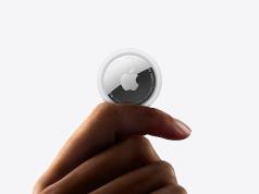 ¿Apple trabajando en un pin de IA? Un movimiento que podría ser una mala decisión estratégica Primer plano de una mano sosteniendo un dispositivo Apple AirTag entre los dedos, con el logotipo de Apple visible, sobre un fondo blanco minimalista.