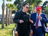 La inminente batalla entre las redes sociales y el estado Un hombre con chaqueta de cuero y gafas de sol conversa con otro hombre vestido con traje azul, corbata roja y una gorra roja que dice "Make America Great Again". Ambos caminan al aire libre en un área ajardinada con palmeras y césped, bajo un cielo despejado.