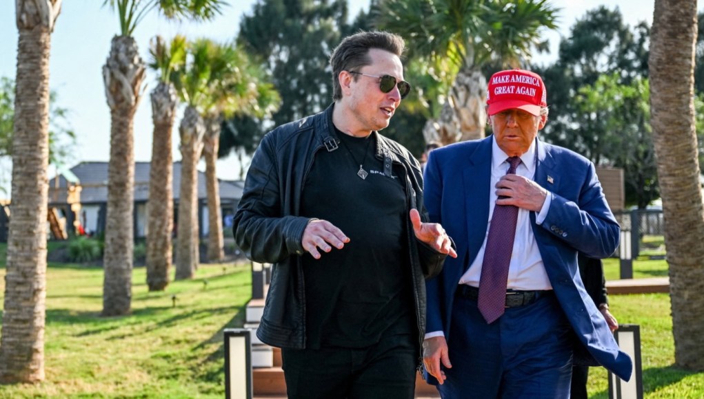 Un hombre con chaqueta de cuero y gafas de sol conversa con otro hombre vestido con traje azul, corbata roja y una gorra roja que dice "Make America Great Again". Ambos caminan al aire libre en un área ajardinada con palmeras y césped, bajo un cielo despejado. Un hombre con chaqueta de cuero y gafas de sol conversa con otro hombre vestido con traje azul, corbata roja y una gorra roja que dice "Make America Great Again". Ambos caminan al aire libre en un área ajardinada con palmeras y césped, bajo un cielo despejado.