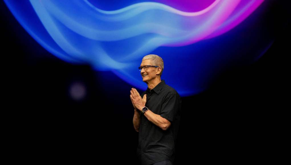 Tim Cook, CEO de Apple, aparece en el escenario durante un evento de lanzamiento de la compañía. Está vestido con una camisa negra y lleva puesto un Apple Watch. En el fondo, se observa una proyección de colores vibrantes en tonos azul y púrpura, creando un efecto de ondas luminosas. Cook sonríe mientras junta las manos, en un gesto de agradecimiento o bienvenida. Tim Cook, CEO de Apple, aparece en el escenario durante un evento de lanzamiento de la compañía. Está vestido con una camisa negra y lleva puesto un Apple Watch. En el fondo, se observa una proyección de colores vibrantes en tonos azul y púrpura, creando un efecto de ondas luminosas. Cook sonríe mientras junta las manos, en un gesto de agradecimiento o bienvenida.