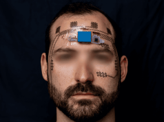 El revolucionario tatuaje electrónico que podría salvar vidas monitoreando el estrés mental de trabajadores Primer plano de un hombre con un dispositivo electrónico experimental adherido a su frente, conectado mediante sensores flexibles que se extienden por el rostro, en un entorno de fondo negro.