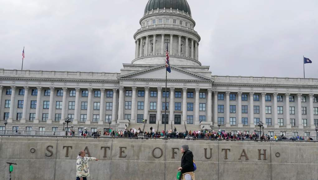 Personas caminando y usando patinetes eléctricos frente al Capitolio del Estado de Utah, un edificio de arquitectura clásica con cúpula, en un día nublado. En primer plano, se lee "STATE OF UTAH" en una pared de cemento. Personas caminando y usando patinetes eléctricos frente al Capitolio del Estado de Utah, un edificio de arquitectura clásica con cúpula, en un día nublado. En primer plano, se lee "STATE OF UTAH" en una pared de cemento.