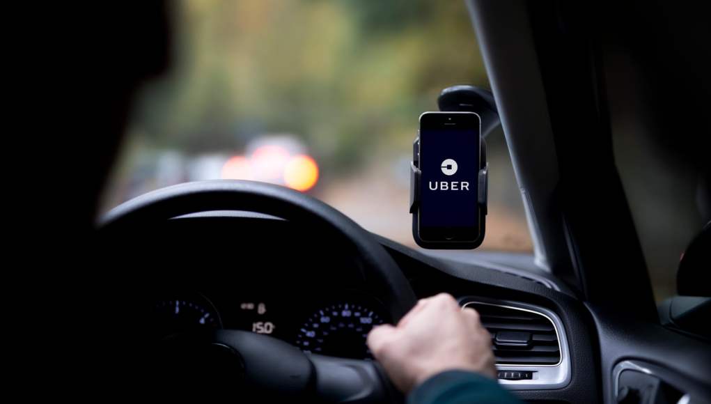 Conductor al volante de un coche, con un smartphone que muestra el logo de Uber fijado al salpicadero. Conductor al volante de un coche, con un smartphone que muestra el logo de Uber fijado al salpicadero.
