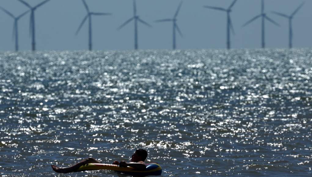Un hombre flota en un inflable sobre el agua en un día soleado, mientras en el horizonte se observan varias turbinas eólicas marinas girando en el mar. El agua brilla intensamente por los reflejos del sol, creando un contraste con las siluetas de las turbinas en el fondo. Un hombre flota en un inflable sobre el agua en un día soleado, mientras en el horizonte se observan varias turbinas eólicas marinas girando en el mar. El agua brilla intensamente por los reflejos del sol, creando un contraste con las siluetas de las turbinas en el fondo.