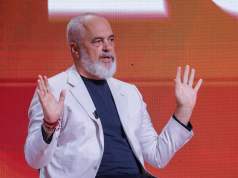 Albania nombra primera ministra de IA para combatir la corrupción en contratos públicos Hombre de cabello canoso y barba blanca, vestido con chaqueta clara y camiseta oscura, sentado frente a un fondo rojo anaranjado. Tiene ambas manos levantadas a la altura del pecho en un gesto expresivo mientras habla.