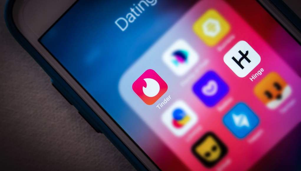 Primer plano de la pantalla de un teléfono inteligente mostrando una carpeta de aplicaciones de citas bajo el nombre 'Dating'. Destacan los iconos de Tinder y Hinge en el centro, junto con otras aplicaciones de citas con colores vibrantes. El fondo de la pantalla tiene un degradado en tonos azul y rosa, mientras que la carcasa del teléfono es azul. Primer plano de la pantalla de un teléfono inteligente mostrando una carpeta de aplicaciones de citas bajo el nombre 'Dating'. Destacan los iconos de Tinder y Hinge en el centro, junto con otras aplicaciones de citas con colores vibrantes. El fondo de la pantalla tiene un degradado en tonos azul y rosa, mientras que la carcasa del teléfono es azul.