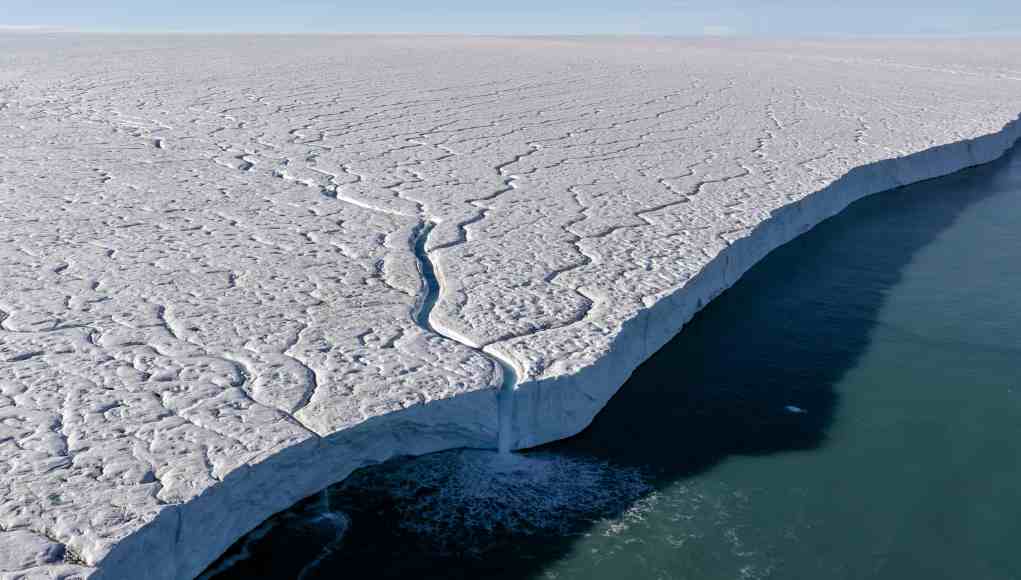 Vista aérea de una gran masa de hielo polar con una grieta por la que fluye agua hacia el océano, destacando los efectos del calentamiento global sobre los glaciares. Vista aérea de una gran masa de hielo polar con una grieta por la que fluye agua hacia el océano, destacando los efectos del calentamiento global sobre los glaciares.