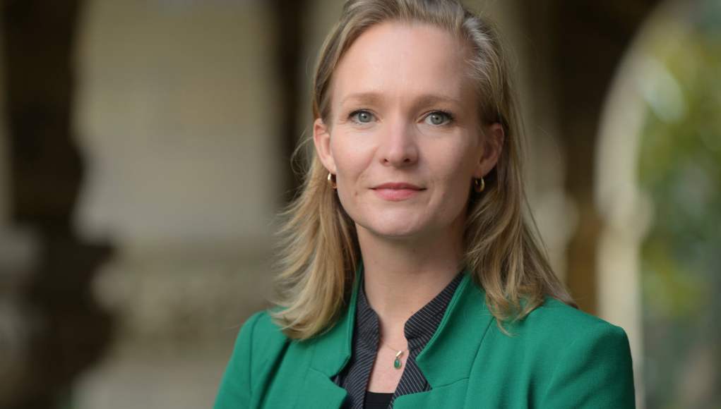 Retrato de una mujer de cabello rubio, con una chaqueta verde y una camisa negra a rayas. Lleva pendientes dorados y un collar con un colgante verde. La imagen muestra un fondo desenfocado con columnas y arcos, lo que sugiere un entorno institucional o académico. Retrato de una mujer de cabello rubio, con una chaqueta verde y una camisa negra a rayas. Lleva pendientes dorados y un collar con un colgante verde. La imagen muestra un fondo desenfocado con columnas y arcos, lo que sugiere un entorno institucional o académico.