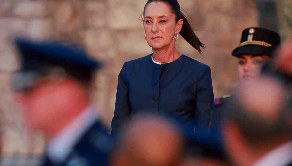 Mujer con expresión seria y vestimenta formal oscura durante un acto oficial, rodeada por personal militar desenfocado en primer plano. Mujer con expresión seria y vestimenta formal oscura durante un acto oficial, rodeada por personal militar desenfocado en primer plano.