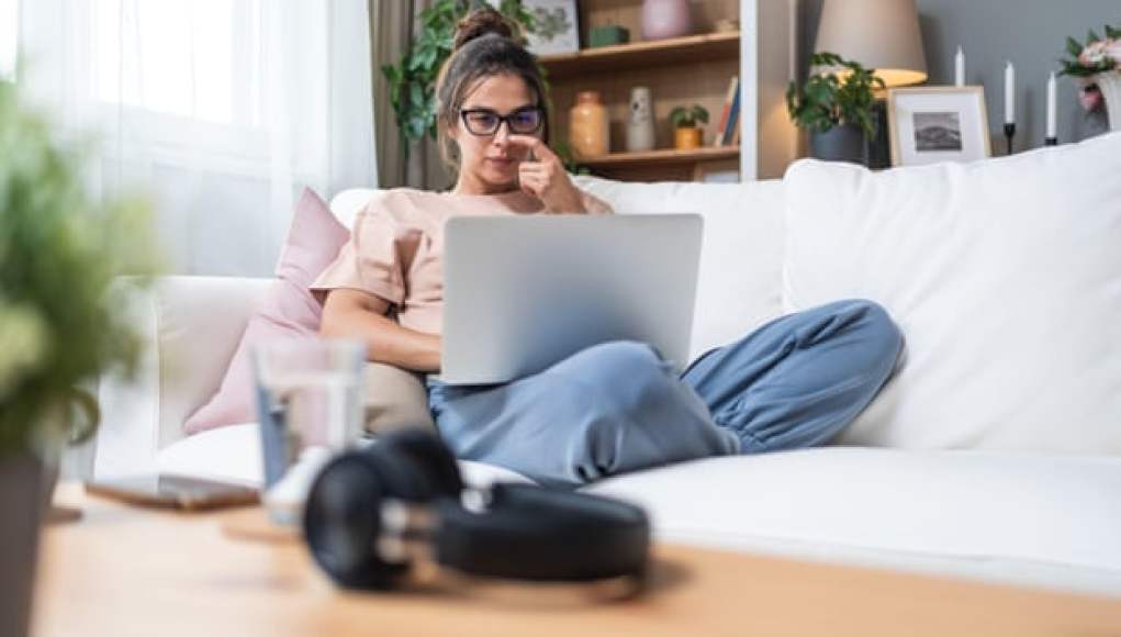 Mujer trabajando desde casa sentada en un sofá, usando un portátil. Lleva gafas y ropa cómoda, rodeada de una decoración hogareña con plantas, estanterías y cojines. En la mesa frente a ella hay unos auriculares, un vaso de agua y otros objetos personales, destacando un ambiente de teletrabajo relajado. Mujer trabajando desde casa sentada en un sofá, usando un portátil. Lleva gafas y ropa cómoda, rodeada de una decoración hogareña con plantas, estanterías y cojines. En la mesa frente a ella hay unos auriculares, un vaso de agua y otros objetos personales, destacando un ambiente de teletrabajo relajado.