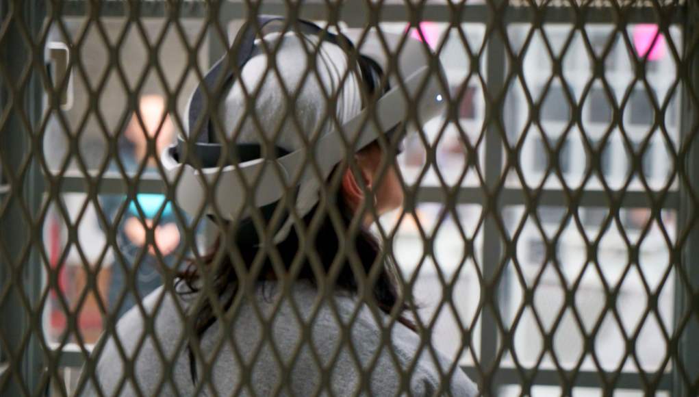 Vista cercana de una persona con un visor de realidad virtual en la cabeza, observada a través de la reja de una celda de malla metálica. Su cabello oscuro es visible debajo del dispositivo. En el fondo, hay otra persona borrosa dentro de una celda similar. La imagen resalta la sensación de confinamiento y el uso de la tecnología en este espacio. Vista cercana de una persona con un visor de realidad virtual en la cabeza, observada a través de la reja de una celda de malla metálica. Su cabello oscuro es visible debajo del dispositivo. En el fondo, hay otra persona borrosa dentro de una celda similar. La imagen resalta la sensación de confinamiento y el uso de la tecnología en este espacio.