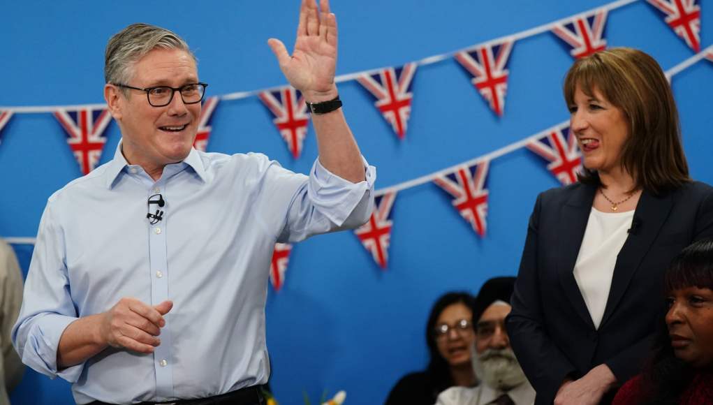 Hombre con camisa clara levanta la mano saludando durante un acto público en un espacio interior decorado con banderines del Reino Unido, mientras una mujer a su lado observa y varias personas escuchan sentadas al fondo. Hombre con camisa clara levanta la mano saludando durante un acto público en un espacio interior decorado con banderines del Reino Unido, mientras una mujer a su lado observa y varias personas escuchan sentadas al fondo.