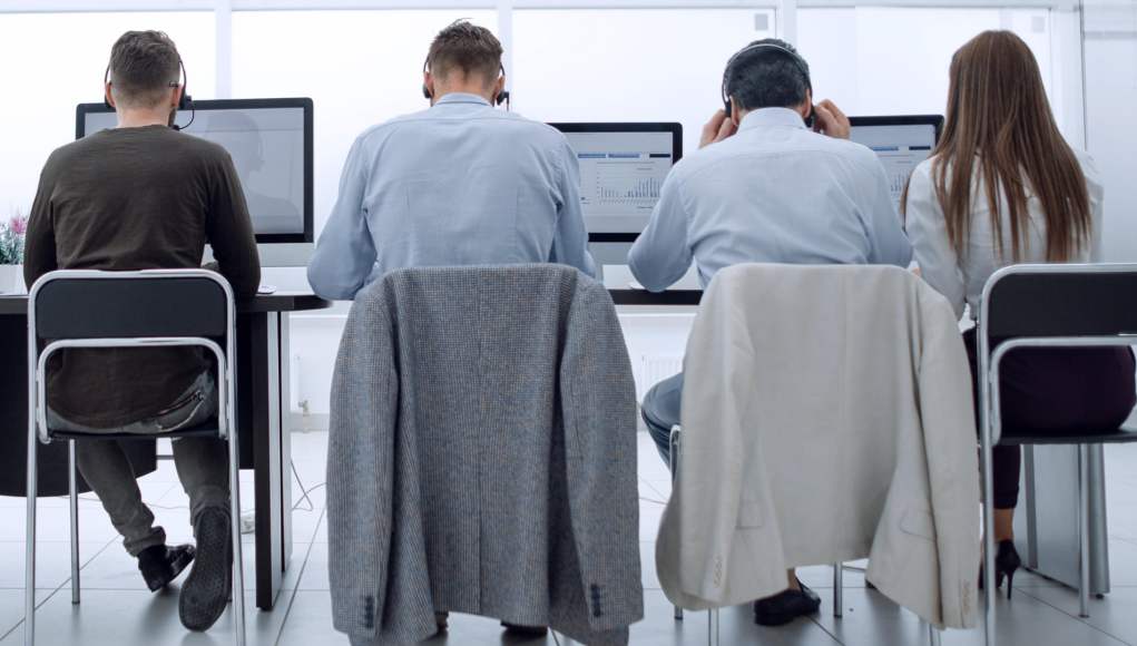 Vista trasera de cuatro personas sentadas frente a ordenadores en una oficina moderna. Llevan auriculares y se concentran en sus pantallas. Dos chaquetas están colgadas en los respaldos de las sillas, lo que sugiere un ambiente profesional o de centro de atención al cliente. Vista trasera de cuatro personas sentadas frente a ordenadores en una oficina moderna. Llevan auriculares y se concentran en sus pantallas. Dos chaquetas están colgadas en los respaldos de las sillas, lo que sugiere un ambiente profesional o de centro de atención al cliente.