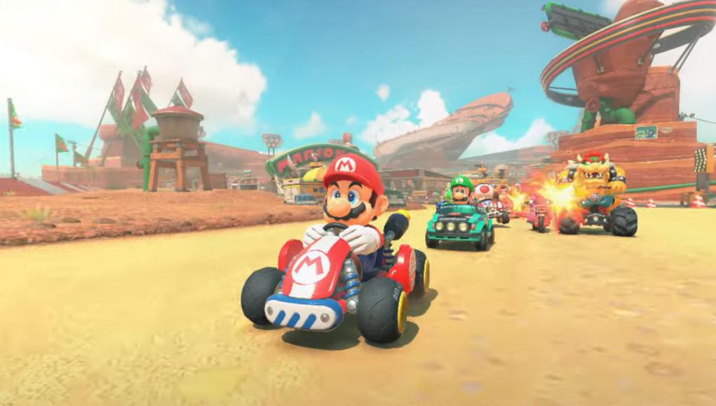 Escena colorida del videojuego Mario Kart con Mario conduciendo un kart rojo en primer plano, seguido por Luigi, Toad y Bowser en una carrera por un circuito ambientado en un paisaje desértico con estructuras y banderas al fondo. Escena colorida del videojuego Mario Kart con Mario conduciendo un kart rojo en primer plano, seguido por Luigi, Toad y Bowser en una carrera por un circuito ambientado en un paisaje desértico con estructuras y banderas al fondo.