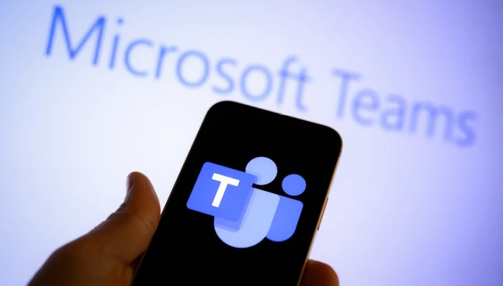 Primer plano de una mano sosteniendo un teléfono móvil con el logotipo de Microsoft Teams en la pantalla, frente a una pantalla de fondo desenfocada que muestra el texto "Microsoft Teams". Primer plano de una mano sosteniendo un teléfono móvil con el logotipo de Microsoft Teams en la pantalla, frente a una pantalla de fondo desenfocada que muestra el texto "Microsoft Teams".