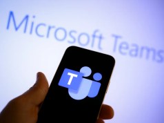 Microsoft intenta esquivar multas de la UE desvinculando Teams y Office Primer plano de una mano sosteniendo un teléfono móvil con el logotipo de Microsoft Teams en la pantalla, frente a una pantalla de fondo desenfocada que muestra el texto "Microsoft Teams".