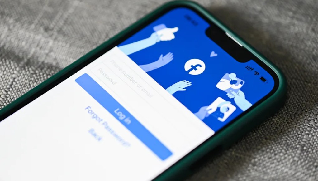 Imagen de un teléfono móvil que muestra la pantalla de inicio de sesión de Facebook, con opciones para ingresar el número de teléfono o correo electrónico y la contraseña. El fondo de la pantalla incluye ilustraciones de manos interactuando con los iconos de "me gusta" y "compartir" de la red social. El dispositivo tiene un borde verde y está sobre una superficie gris. Imagen de un teléfono móvil que muestra la pantalla de inicio de sesión de Facebook, con opciones para ingresar el número de teléfono o correo electrónico y la contraseña. El fondo de la pantalla incluye ilustraciones de manos interactuando con los iconos de "me gusta" y "compartir" de la red social. El dispositivo tiene un borde verde y está sobre una superficie gris.