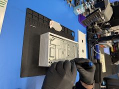 Microsoft y iFixit ofrecen piezas de repuesto oficiales para Xbox Series X/S para reparaciones DIY Un técnico con guantes negros utiliza una herramienta para reparar una consola Xbox Series S desmontada, que está sobre una superficie de trabajo con un tapete negro. En el fondo, se observan herramientas adicionales y equipos electrónicos organizados en el espacio de trabajo.