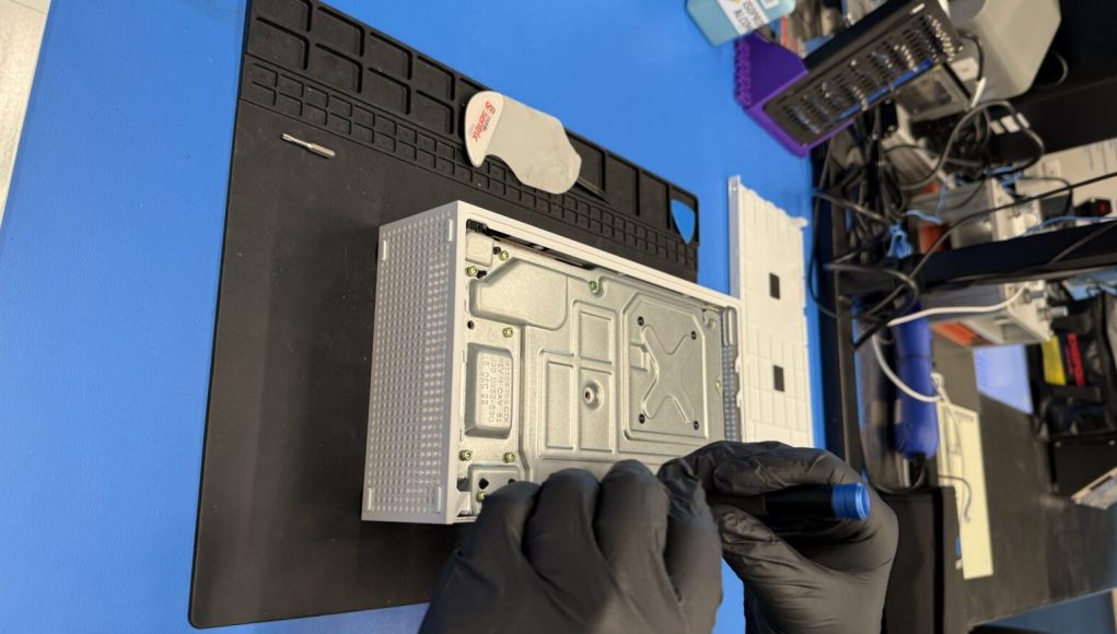 Un técnico con guantes negros utiliza una herramienta para reparar una consola Xbox Series S desmontada, que está sobre una superficie de trabajo con un tapete negro. En el fondo, se observan herramientas adicionales y equipos electrónicos organizados en el espacio de trabajo. Un técnico con guantes negros utiliza una herramienta para reparar una consola Xbox Series S desmontada, que está sobre una superficie de trabajo con un tapete negro. En el fondo, se observan herramientas adicionales y equipos electrónicos organizados en el espacio de trabajo.