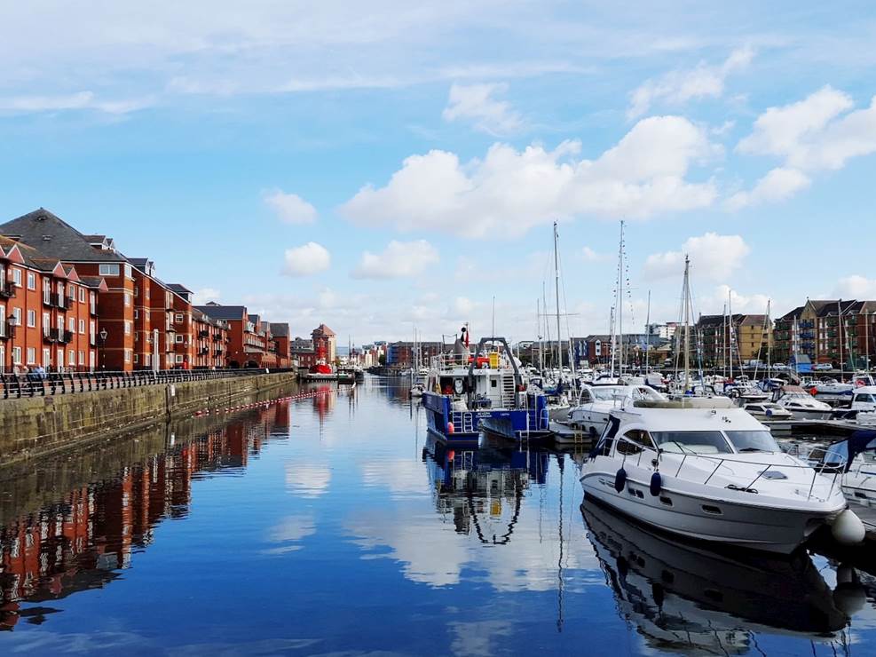 A scenic view of Swansea Marina showcasing boats moored along the waterfront with city buildings in the background. A scenic view of Swansea Marina showcasing boats moored along the waterfront with city buildings in the background.